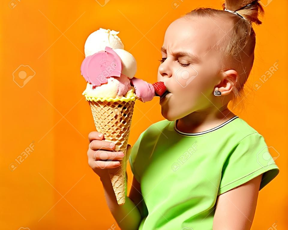 Pretty baby girl kid eating licking banana and strawberry ice cream in waffles cone on yellow background with free text copy space