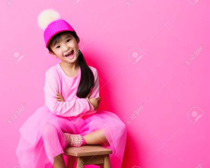 Nicely smiling asian fashion kid girl in pink dress and funny cap with fur pompon on pink background with free text space