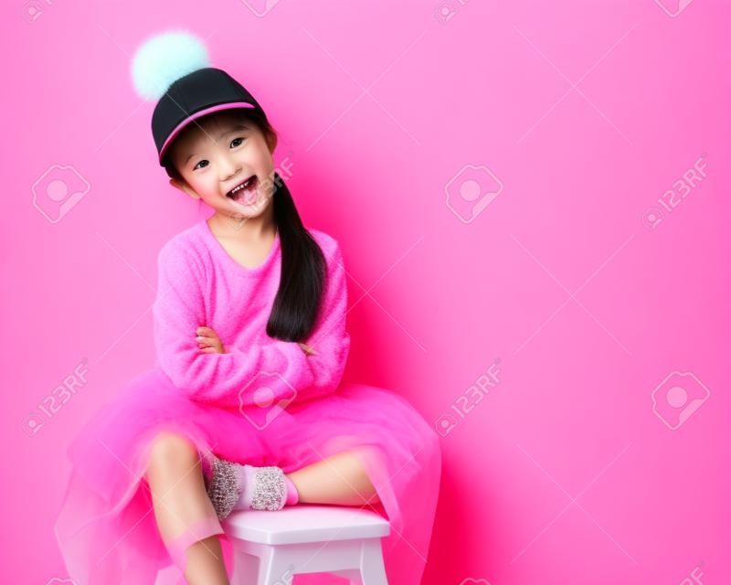 Nicely smiling asian fashion kid girl in pink dress and funny cap with fur pompon on pink background with free text space