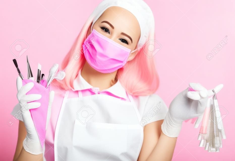 Nail technician in pink medicine gloves, apron and mask with nail varnish samples display and manicure equipment in waffle cone on pink