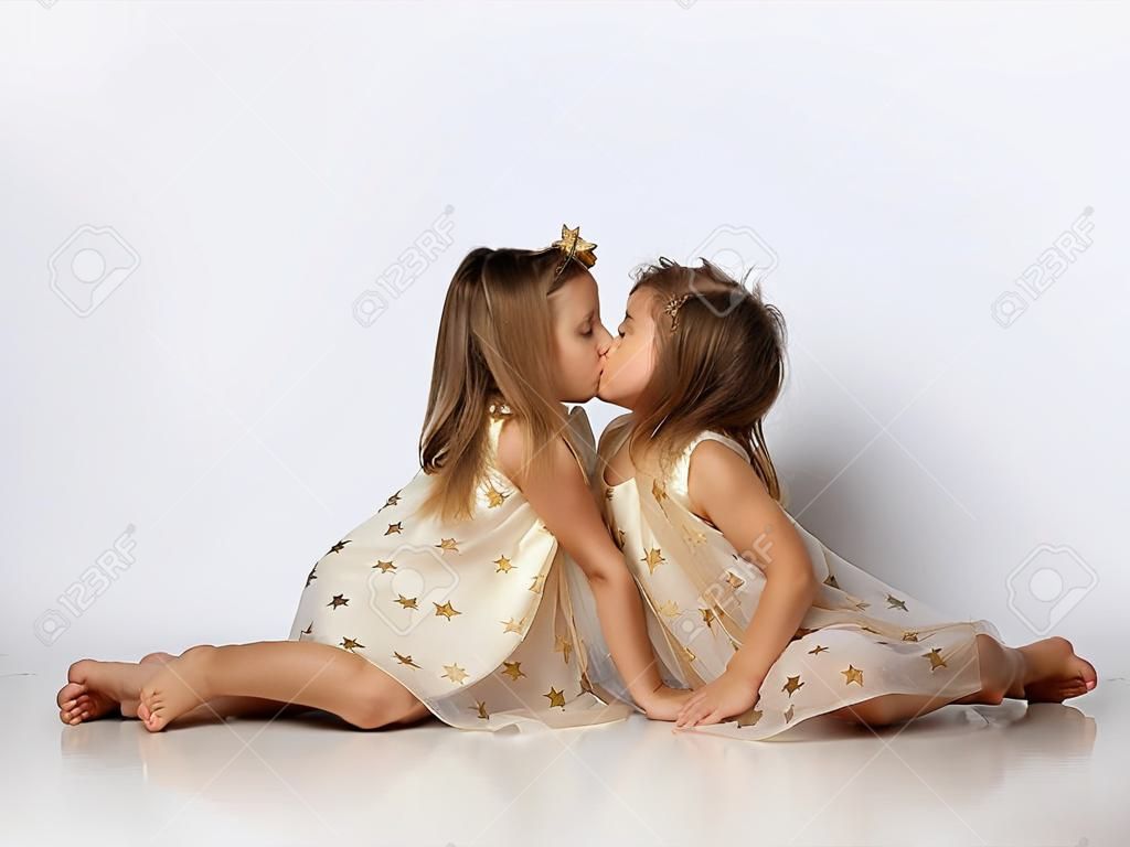 Twee kleine mooie meisjes zussen in dezelfde jurken met sterren blootsvoets zitten op de vloer en elkaar kussen over grijs