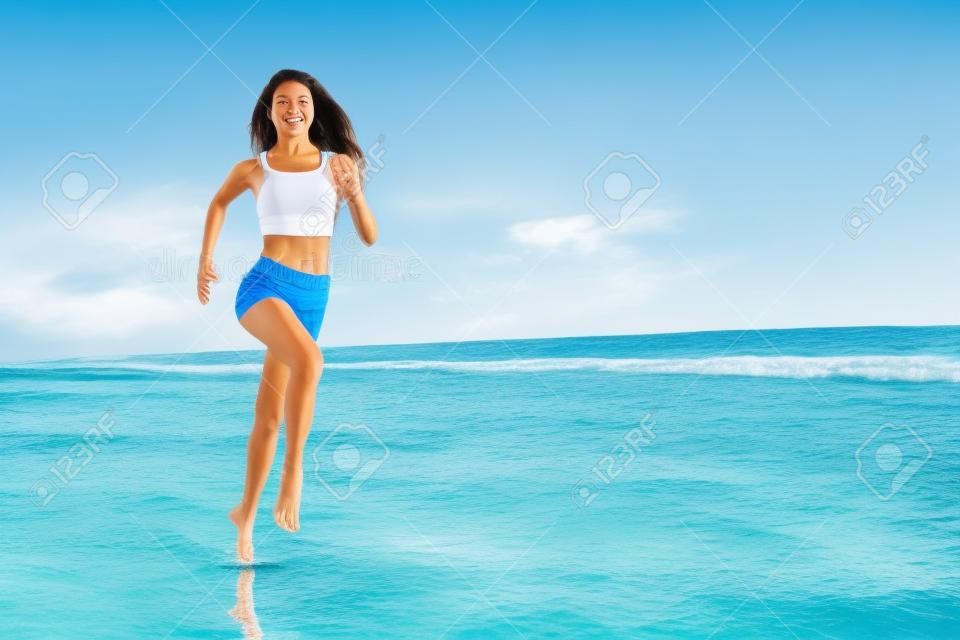 Barefoot young girl with slim body running along sea surf by water pool to keep fit and burning fat. Beach background with blue sky. Woman fitness, jogging sports activity on summer family vacation.