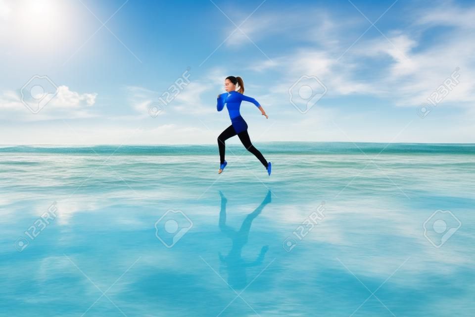 Barefoot young girl with slim body running along sea surf by water pool to keep fit and burning fat. Beach background with blue sky. Woman fitness, jogging sports activity on summer family vacation.