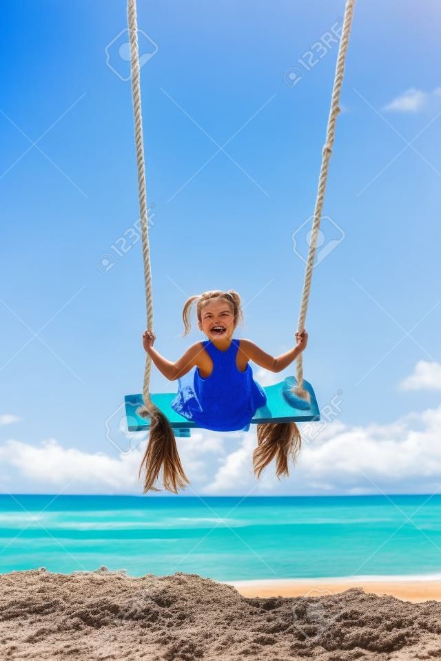 Happy girl have fun swinging high in mid air. Flying up upside down on rope swing on sea beach. Travel adventure on paradise tropical island. Family lifestyle, activity on summer vacation with kids