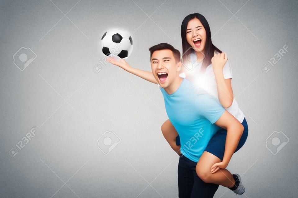 Вдохновленная молодая пара, женщина сидит на спине человека, болельщики с футбольным мячом аплодируют любимой футбольной команде, выразительно жестикулируя руками, изолированными на белом фоне. Семейный досуг, концепция образа жизни