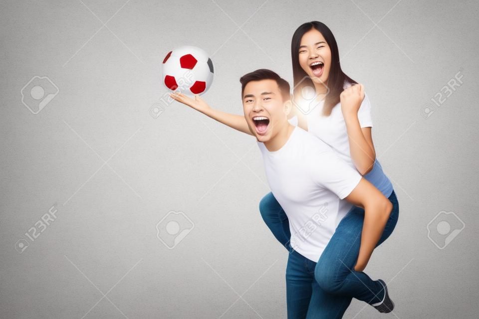Вдохновленная молодая пара, женщина сидит на спине человека, болельщики с футбольным мячом аплодируют любимой футбольной команде, выразительно жестикулируя руками, изолированными на белом фоне. Семейный досуг, концепция образа жизни
