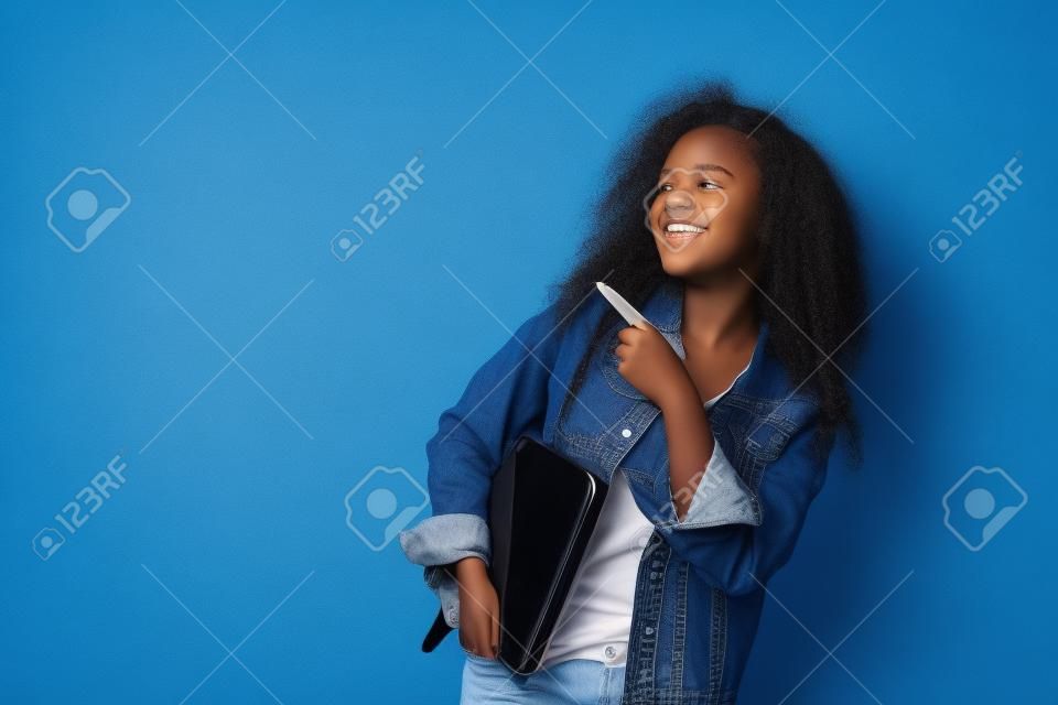 Young african american girl teen student in denim clothes, backpack hold pc isolated on blue wall background studio portrait. Education in high school university college concept. Mock up copy space