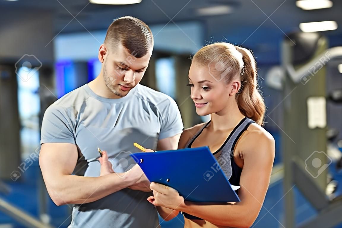 fitness, sport, ćwiczenia i dieta - uśmiecha się młoda kobieta i osobistego trenera ze schowka piśmie plan ćwiczeń w siłowni