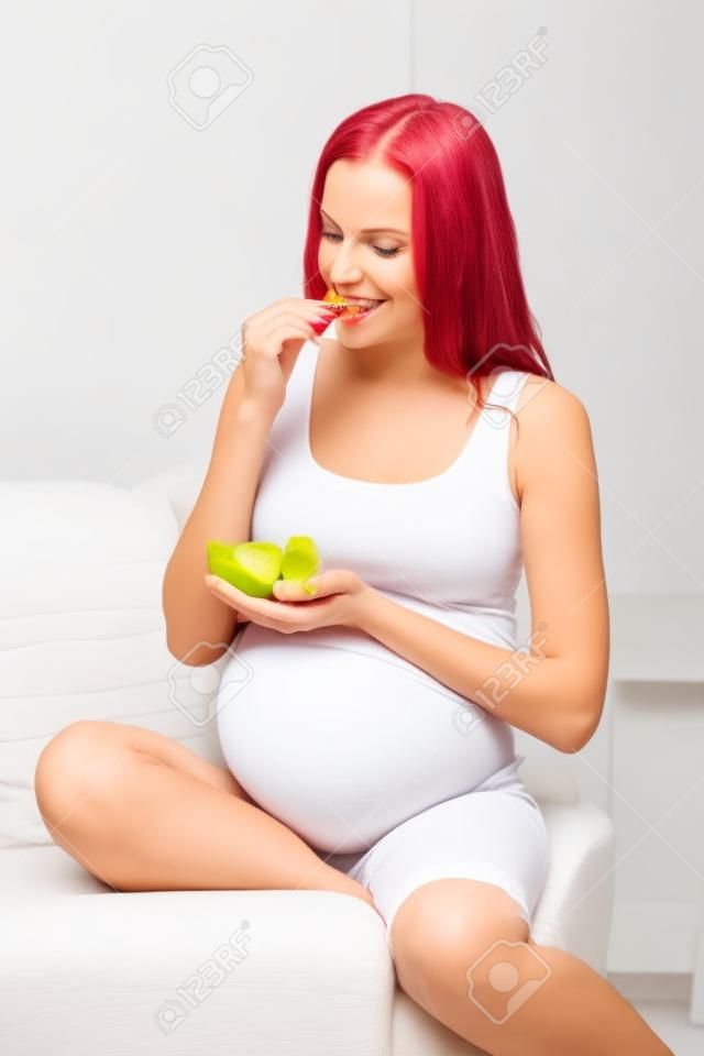 el embarazo, la alimentación, la alimentación saludable, la gente y la expectativa concepto - mujer embarazada feliz comiendo las frutas en el hogar