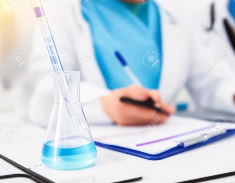 science, chemistry, biology, medicine and people concept - close up of scientist with pipette and flask taking notes to clipboard in clinical laboratory