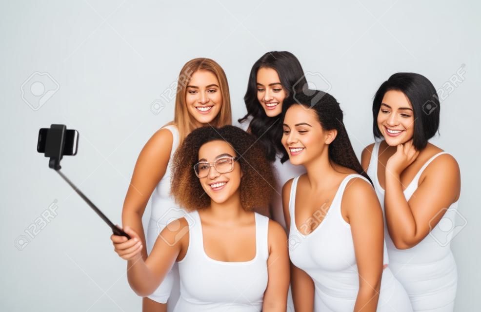 technology, friendship, body positive and people concept - group of happy women in white underwear taking picture with smartphoone on selfie stick