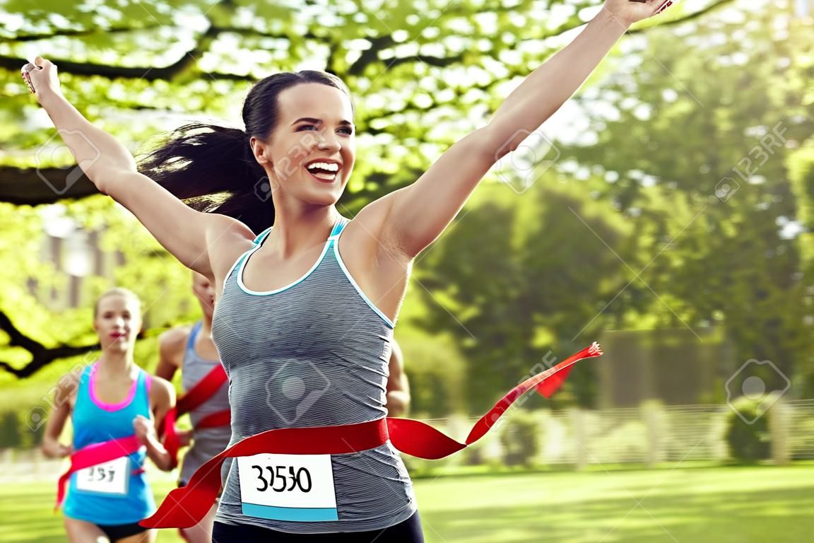 健身运动胜利的成功和健康的生活方式的概念-快乐女子赢得比赛，并率先完成红色丝带超过一群运动员运行马拉松与徽章号码户外