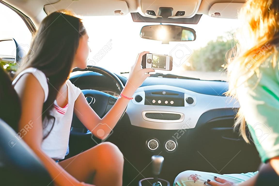 nyári vakáció, nyaralás, utazás, utazásairól és az emberek koncepció - boldog tizenéves lányok vagy fiatal nők okostelefon figyelembe szelfi az autó