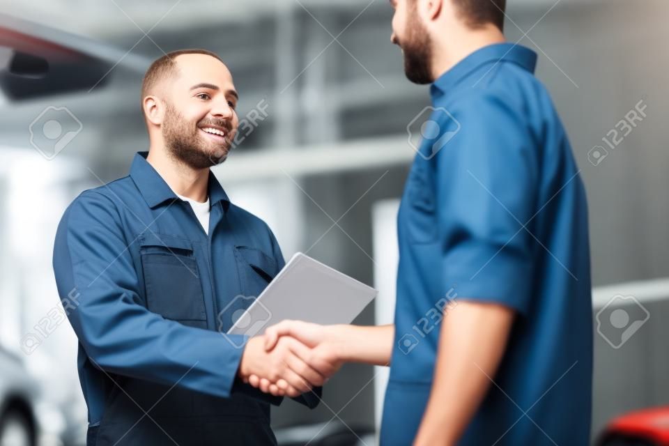 auto service, repair, maintenance, gesture and people concept - mechanic with clipboard and man or owner shaking hands at car shop