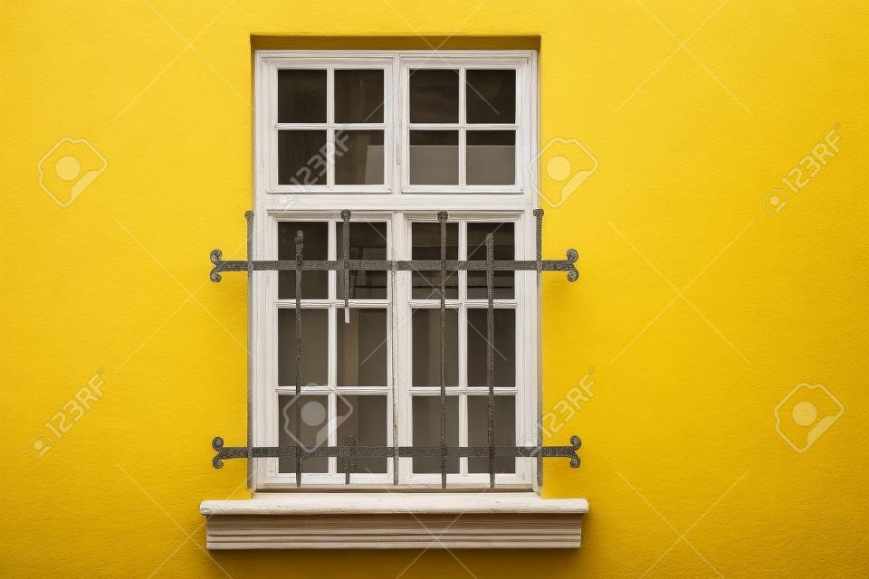 Fenster mit weißem rechteckigem Rahmen und Einfassung, an der gelben Hauswand und geschlossenen dekorativen Eisenstangen. Aus der Serie - Fenster der Welt.