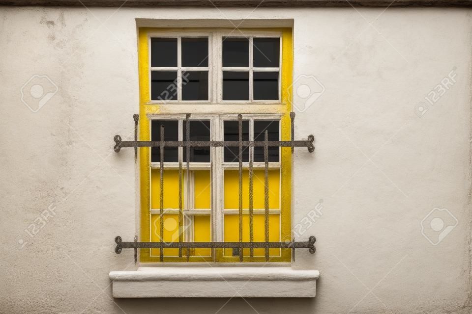 Fenster mit weißem rechteckigem Rahmen und Einfassung, an der gelben Hauswand und geschlossenen dekorativen Eisenstangen. Aus der Serie - Fenster der Welt.