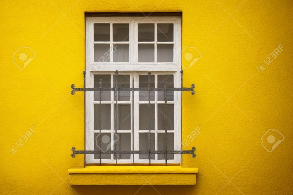 Okno z białą prostokątną ramą i oprawą, usytuowane na żółtej ścianie domu i zamykanymi ozdobnymi żelaznymi kratami. Z serii - Okna świata.