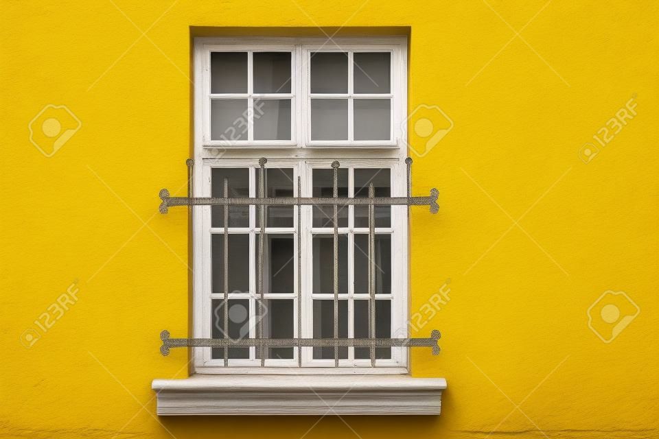 Ventana con marco rectangular blanco y encuadernación, ubicada en la pared amarilla de la casa y rejas de hierro decorativas cerradas. De la serie - Ventanas del mundo.