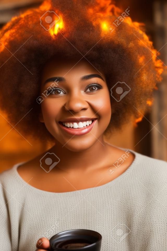 Happy joyful african american woman drinking cup of coffee relaxing at fireplace. Young black girl with hot beverage heating warming up. autumn at home.