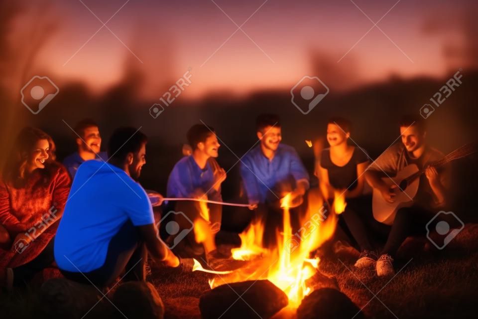 Un gruppo di giovani amici felici che si rilassano e si godono la serata estiva intorno al fuoco sulla riva del fiume