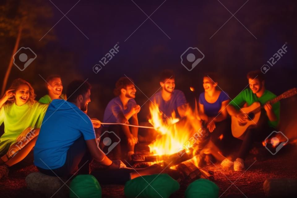 Eine Gruppe glücklicher junger Freunde entspannt sich und genießt den Sommerabend am Lagerfeuer am Flussufer