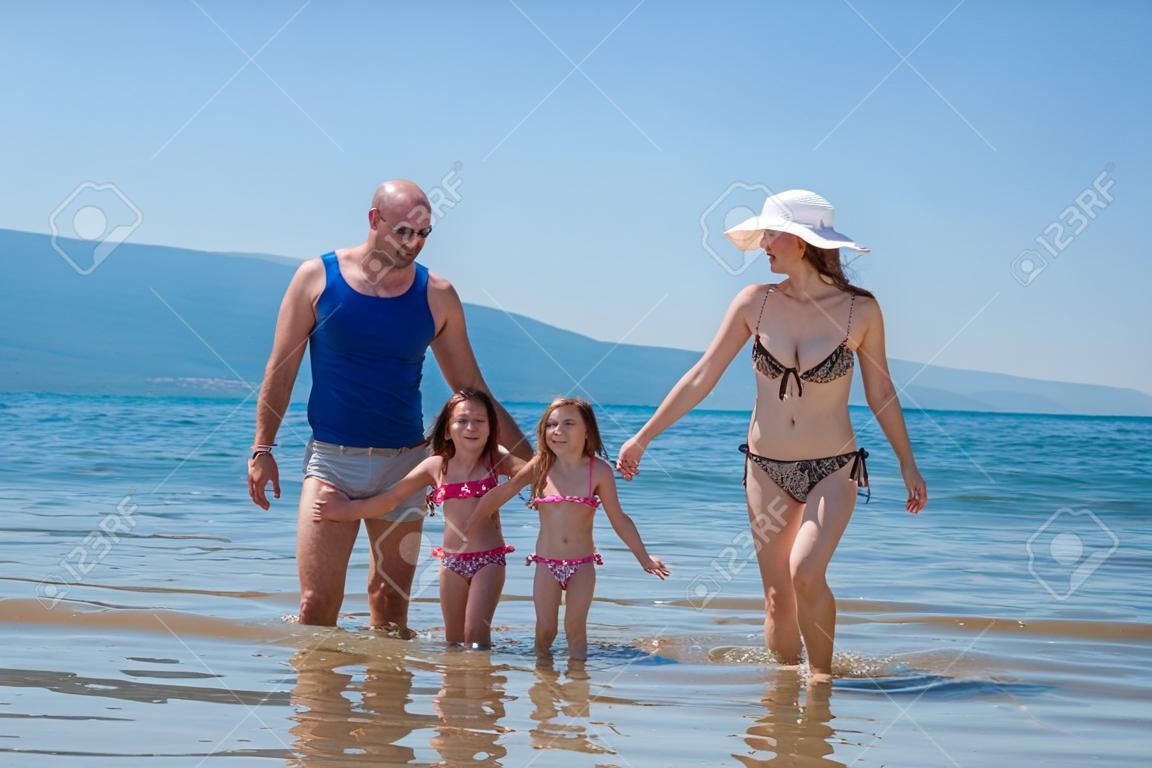 Portrait of a young happy couple with cute little daughters having fun while standing in the shallow water of the sea during Summer vacation  Healthy family holiday concept