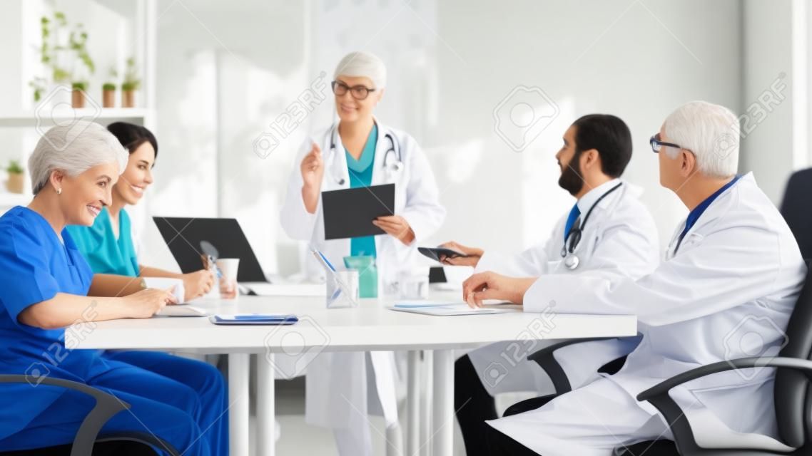 Portret dojrzałej kobiety, lekarza prowadzącego spotkanie komitetu w radzie lekarskiej, przedstawiającej nowe procedury medyczne stojące przy biurku. grupa lekarzy rozmawiających o objawach choroby w biurze kliniki