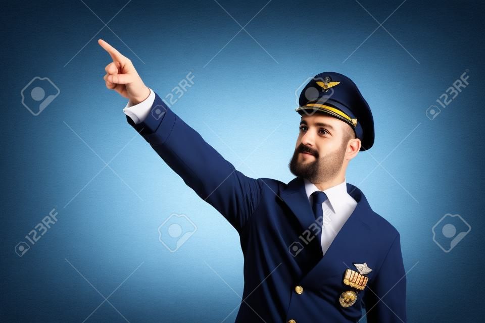 Serious captain pointing at sky with index finger, wearing professional aviation uniform, plane pilot looking at camera. Confident aviation academy aviator standing, studio medium shot