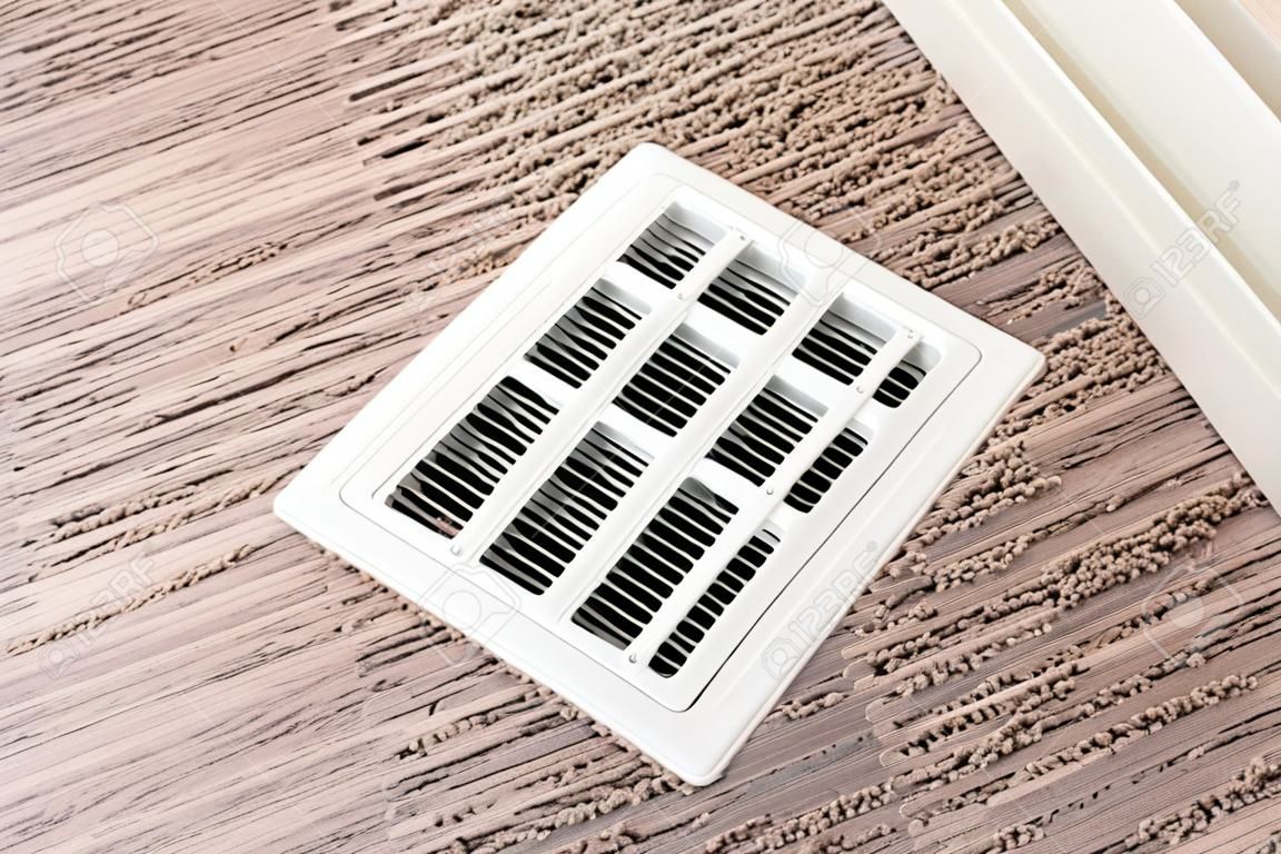 Biała osłona kratki kanału klimatyzatora na podłodze z brązowym dywanem. osłona posiada regulowane szczeliny liniowe umożliwiające skierowanie strumienia powietrza do wnętrza pomieszczenia.
