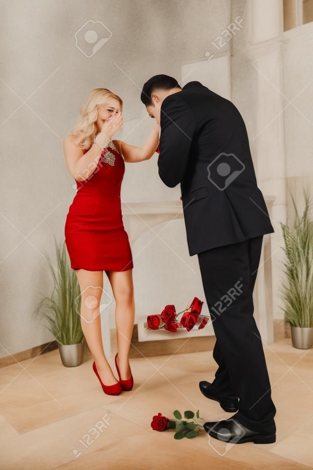 longstemmed 빨간색으로 그녀의 손에 키스하고 몸을 굽히고 아름다운 여자를 제안하는 기사도 젊은 남자가 자신의 발에 누워 장미
