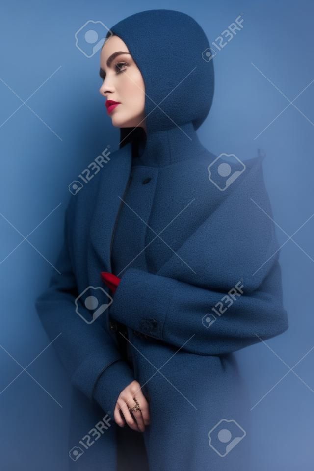 Bella donna in cappotto autunnale blu. Giovane bella posa castana in studio in abiti vintage. Signora alla moda con l'acconciatura classica professionale. Ritratto di arte di moda.