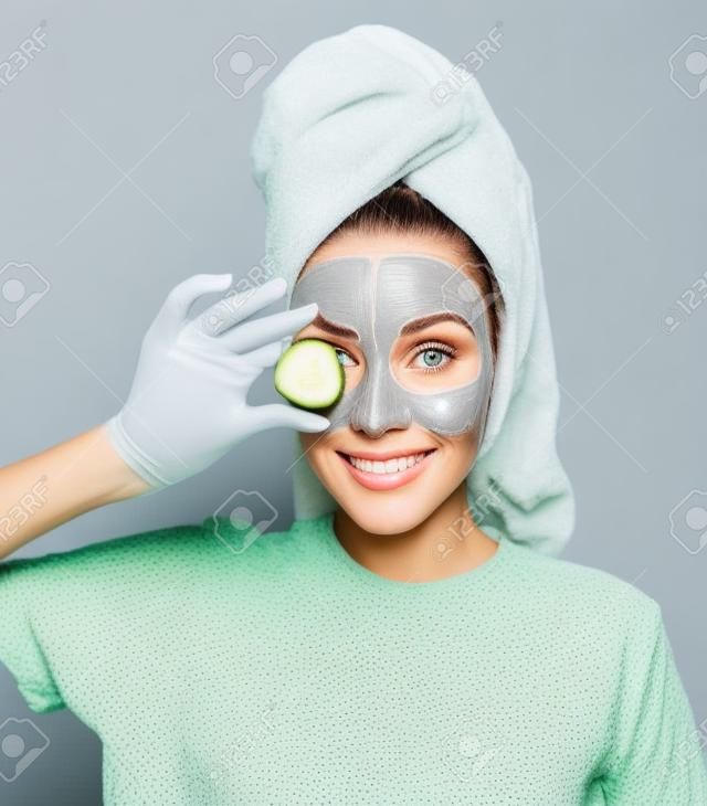 mulher bonita com uma máscara cinza argila em seu rosto segura uma fatia de pepino fresco em sua mão. Cuidados com a pele em casa.