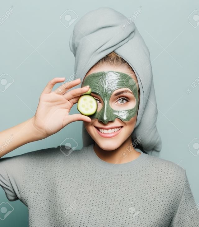 mulher bonita com uma máscara cinza argila em seu rosto segura uma fatia de pepino fresco em sua mão. Cuidados com a pele em casa.