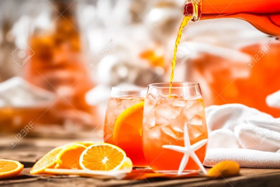 Barman은 오렌지 슬라이스가 있는 두 잔 중 하나에 aperol spritz 칵테일을 붓고 있습니다. 일부 조개껍데기, 수건, 선글라스는 수영장에 닫혀 있습니다. 휴가 개념.