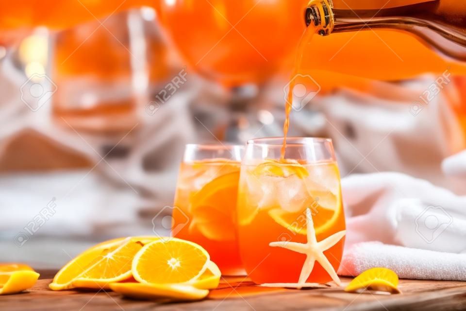 Barman is pouring aperol spritz cocktail into one of two glasses with orange slices. Some seashells, towel and sunglasses closed to swimming pool. Vacation concept.
