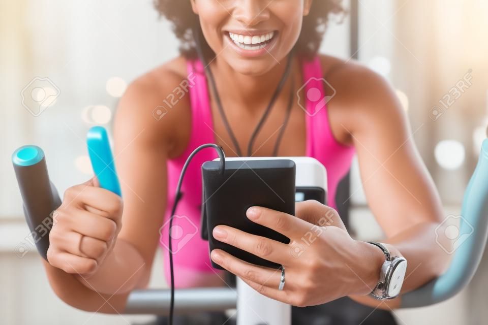 Saudável ajuste sorridente mulher treinando em casa no exercício bicicleta estática durante o treino segurando o telefone e fazendo polegares para cima sinal ouvir música com fones de ouvido para motivação.