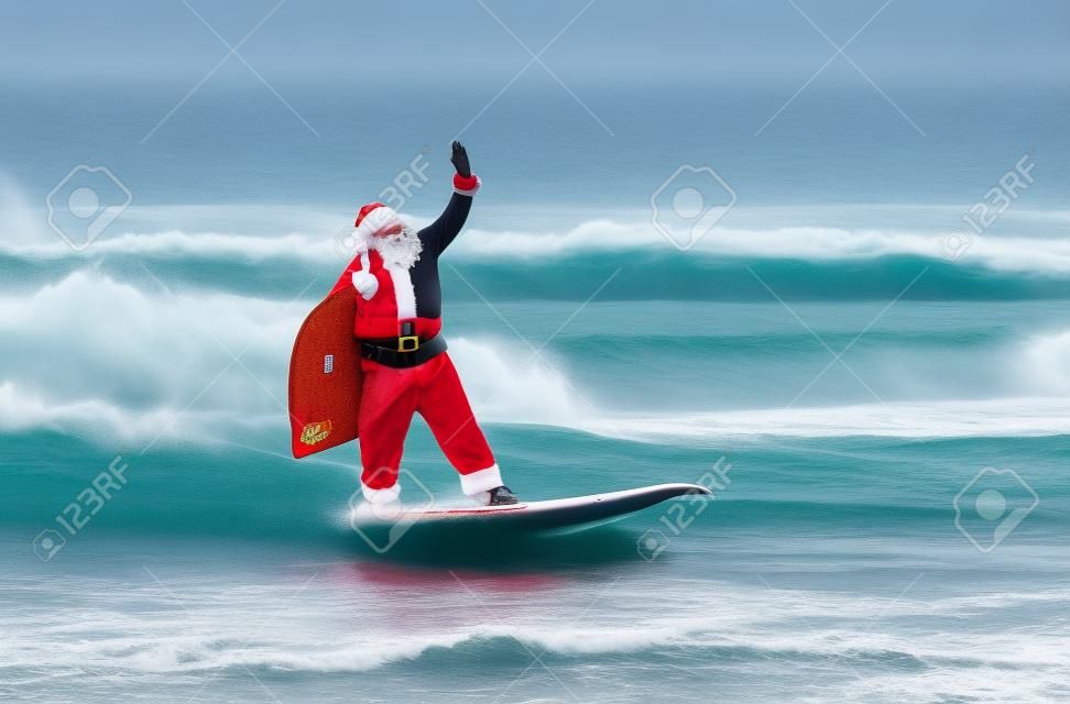 Santa Claus windsurfer with large holiday gifts sack go surfing with surfboard at ocean waves splashes in windy weather - New Year and Christmas active sports lifestyle concept