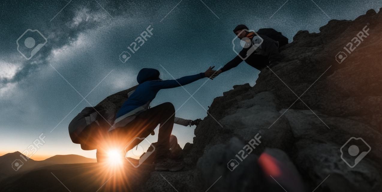Un par de excursionistas ayudándose unos a otros a escalar una montaña al atardecer. Personas que dan una mano amiga y un concepto de deporte activo.