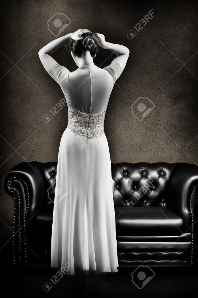 Black and white rear view of elegant woman in evening dress with open back posing at vintage leather couch and textured grunge background