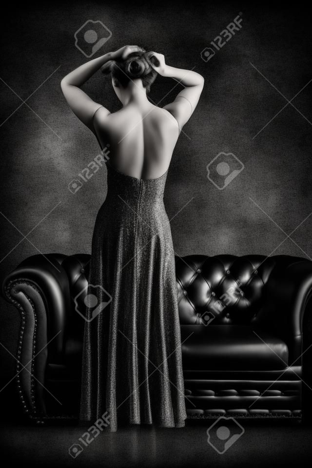 Черно-белый вид сзади элегантная женщина в вечернем платье с открытой спиной, создает в винтажном кожаный диван и текстурированный фон гранж
