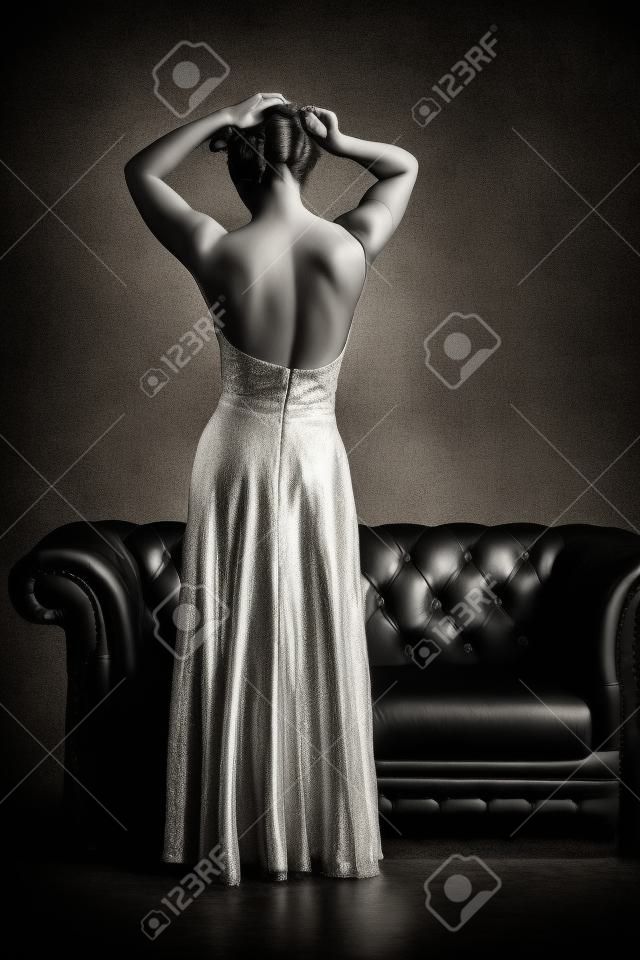 Czarno-biały widok z tyłu kobiety w eleganckiej wieczorowej sukni z otwartym powrotem stwarzających na skórzanej kanapie i rocznika grunge teksturowane