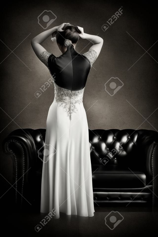 Black and white rear view of elegant woman in evening dress with open back posing at vintage leather couch and textured grunge background