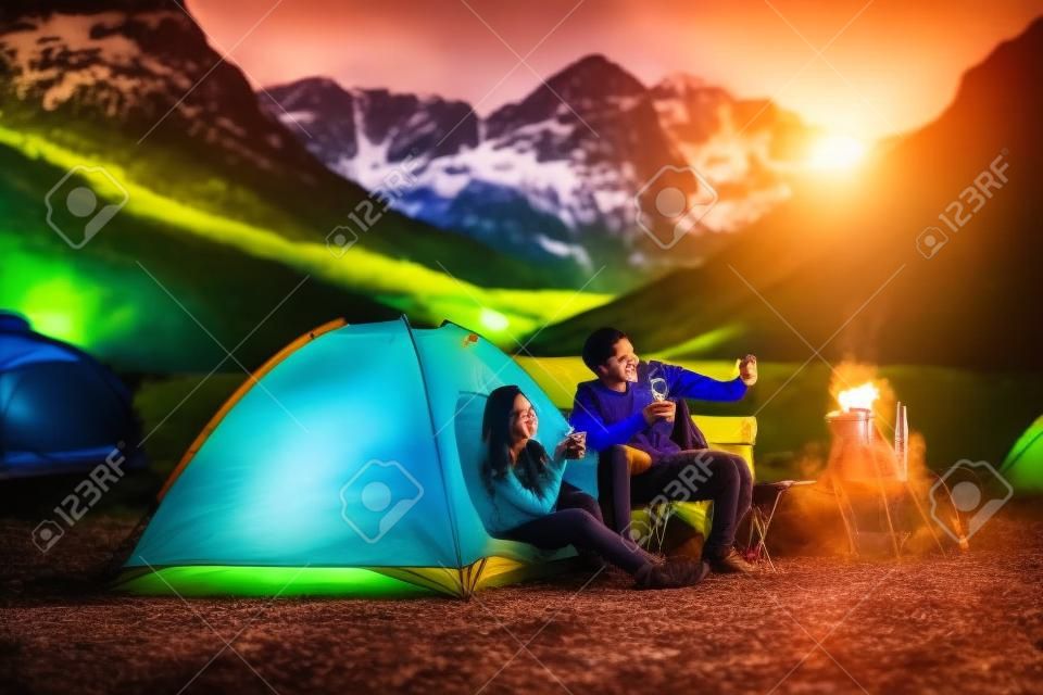 Fröhliches junges asiatisches indisches Paar, das beim Zelten auf dem Campingplatz sitzt, sich entspannt und Tee trinkt.