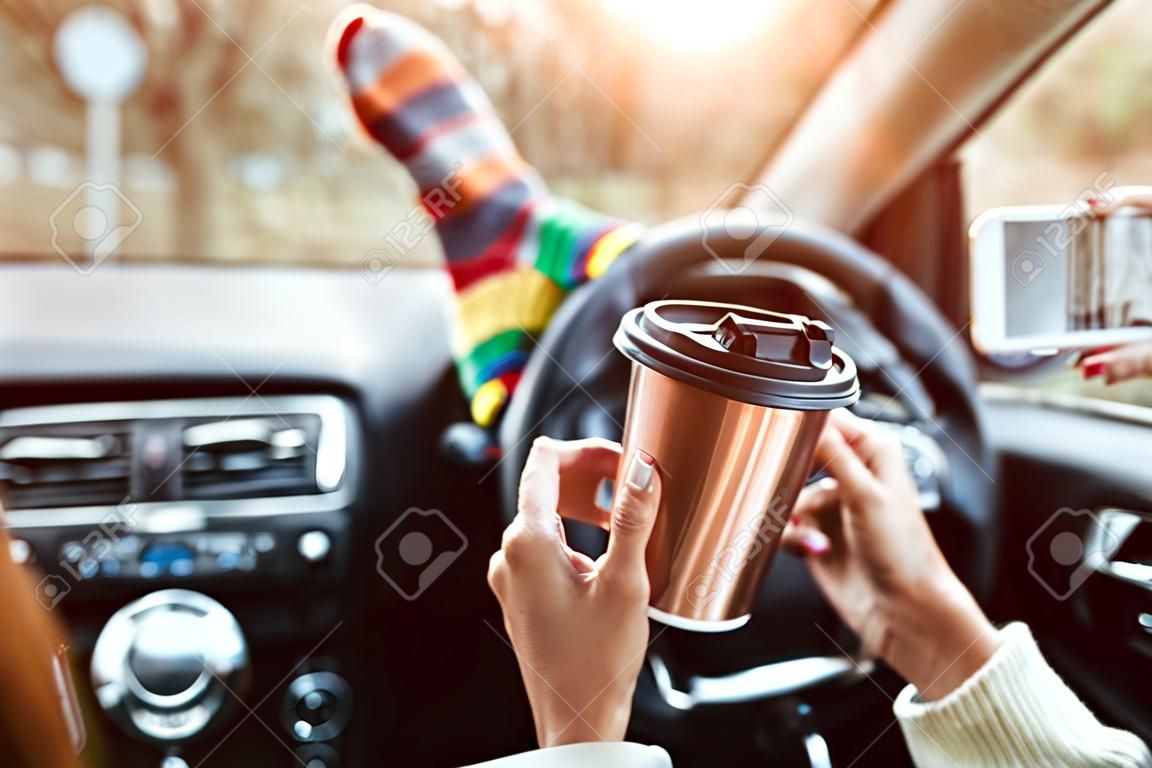 Autunno, viaggio in auto. Cose-up di una donna che beve portare via la tazza di caffè durante il viaggio in auto. Piedi di donna in calzini caldi sul cruscotto dell'auto. Bere portare via il caffè e usare uno smartphone su strada.