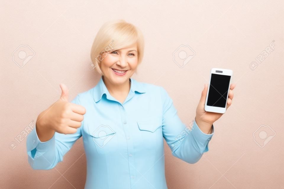 A picture of mature woman with new smartphone. She has tested it and admitted this phone is a good one. Thats why she shows a big thumb up. Isolated on white background.
