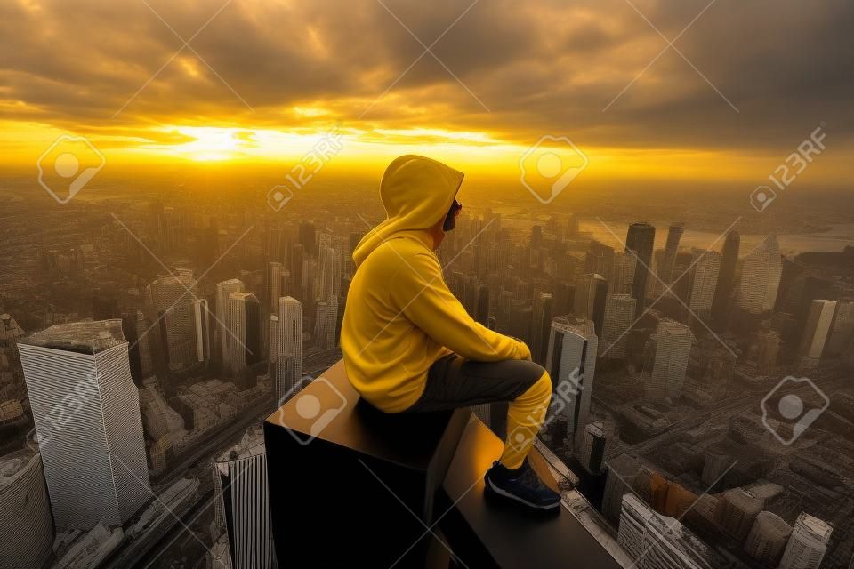 Un hombre con una sudadera con capucha amarilla se sienta en el borde de un rascacielos y mira la puesta de sol