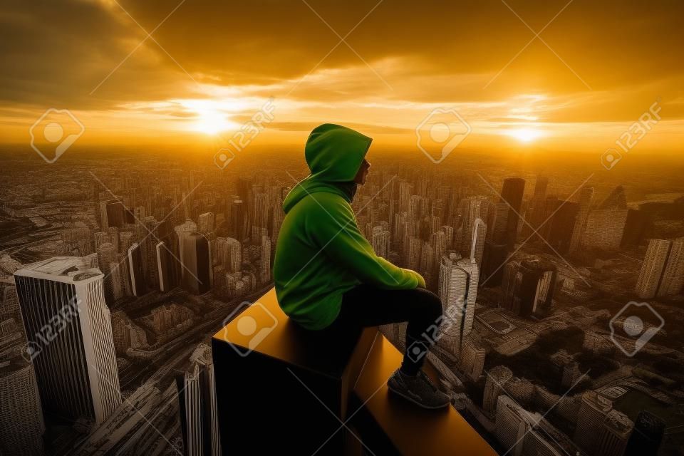 A man in a yellow hooded sweatshirt sits on the edge of a skyscraper and looks at the sunset