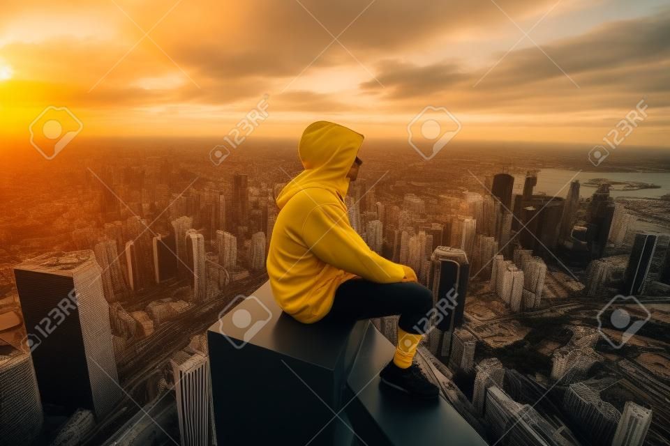 A man in a yellow hooded sweatshirt sits on the edge of a skyscraper and looks at the sunset