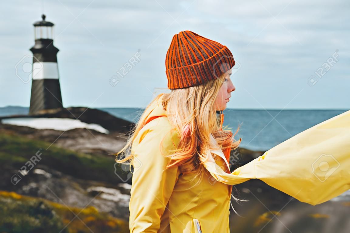 Frauenbesichtigungsleuchtturm-Seelandschaft in den skandinavischen Ferien Norwegen-Reise-Lebensstilkonzeptes im Freien Blondes Mädchenhaar auf dem Wind, der orange Hut und gelben Regenmantel trägt