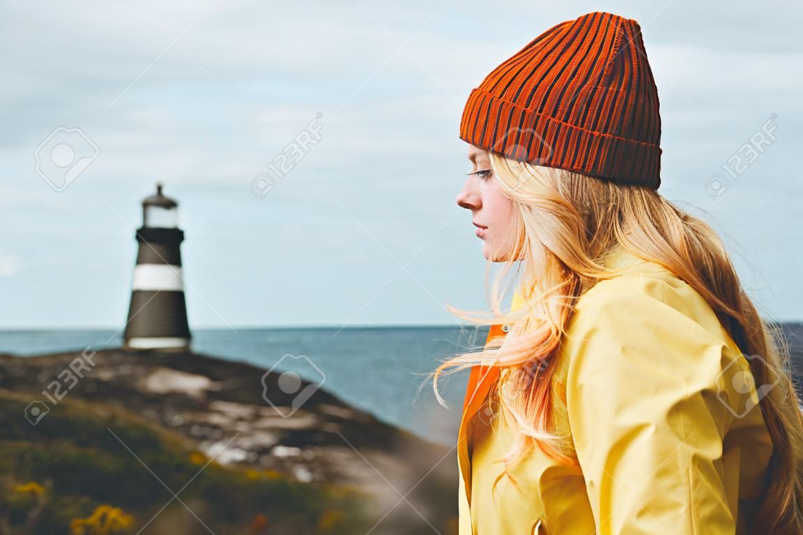 Frauenbesichtigungsleuchtturm-Seelandschaft in den skandinavischen Ferien Norwegen-Reise-Lebensstilkonzeptes im Freien Blondes Mädchenhaar auf dem Wind, der orange Hut und gelben Regenmantel trägt